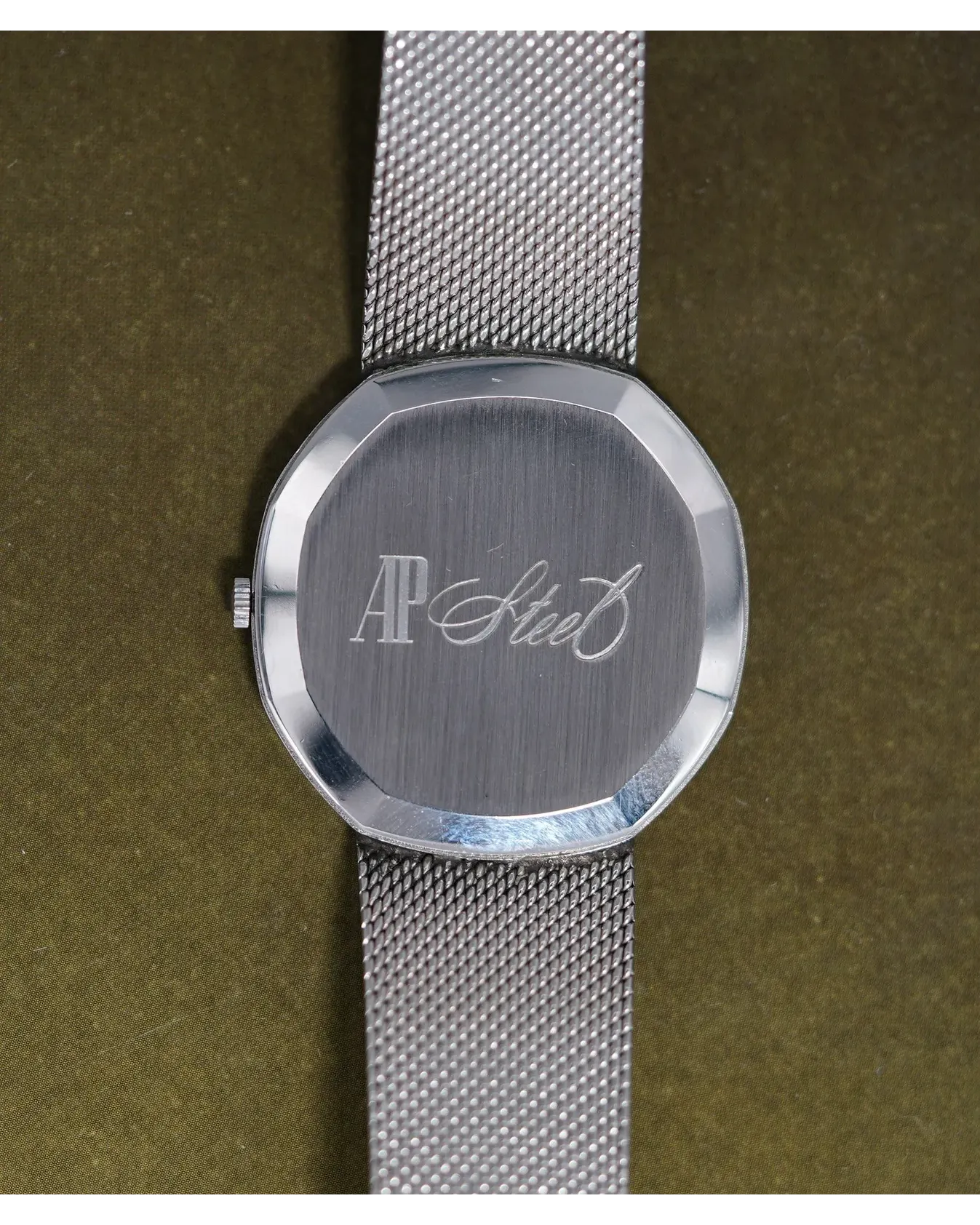 Caseback view of an AP Steel wristwatch with brushed finish and mesh bracelet on olive background.