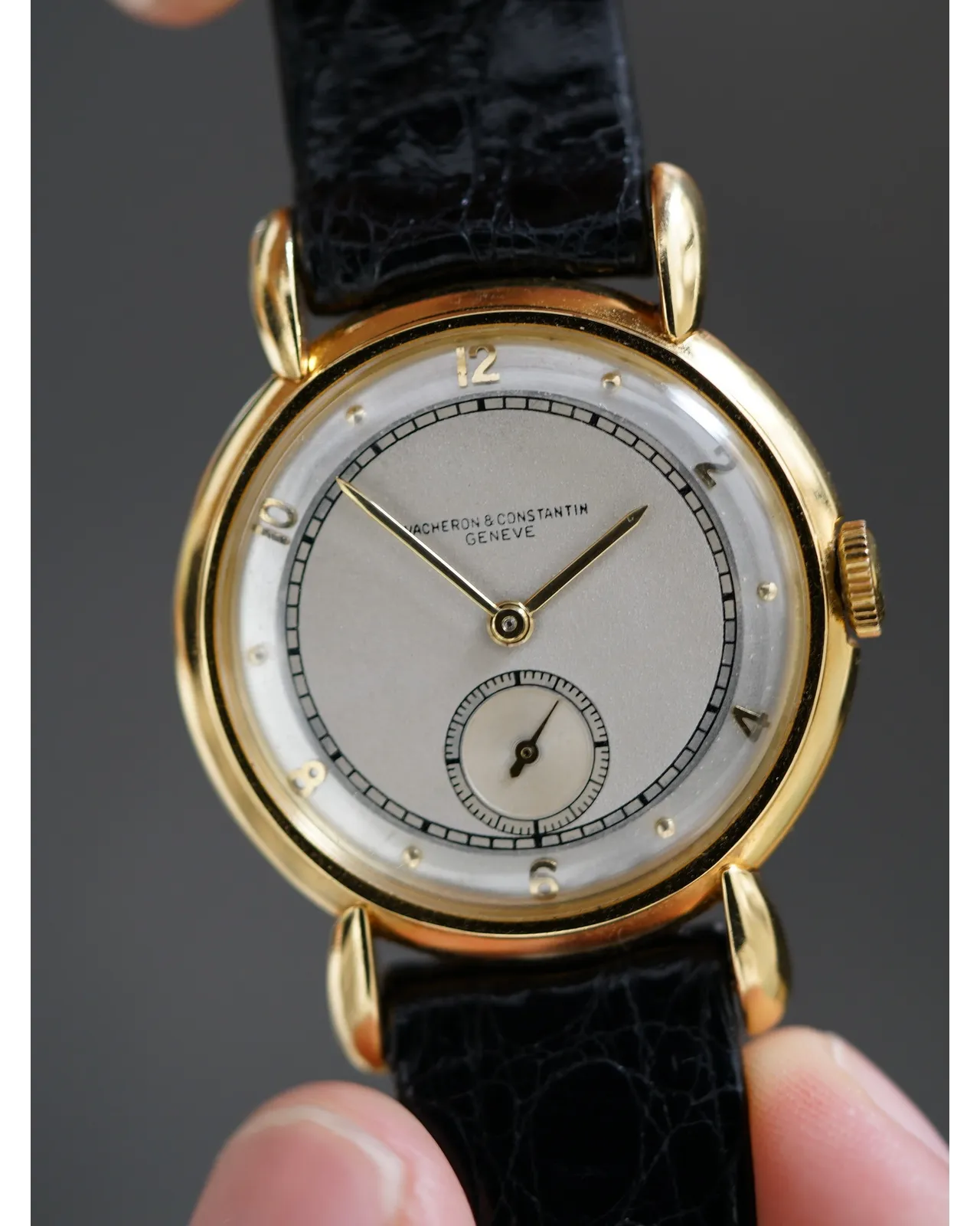 Hands-on shot of a Vacheron Constantin yellow gold watch with two-tone dial, sub-seconds, and black leather strap.