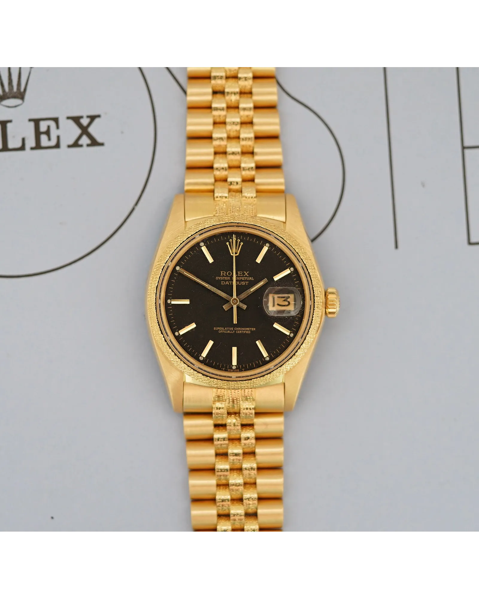 Flat-lay photograph of a yellow gold Rolex Datejust with black dial, baton indices, and Jubilee bracelet.