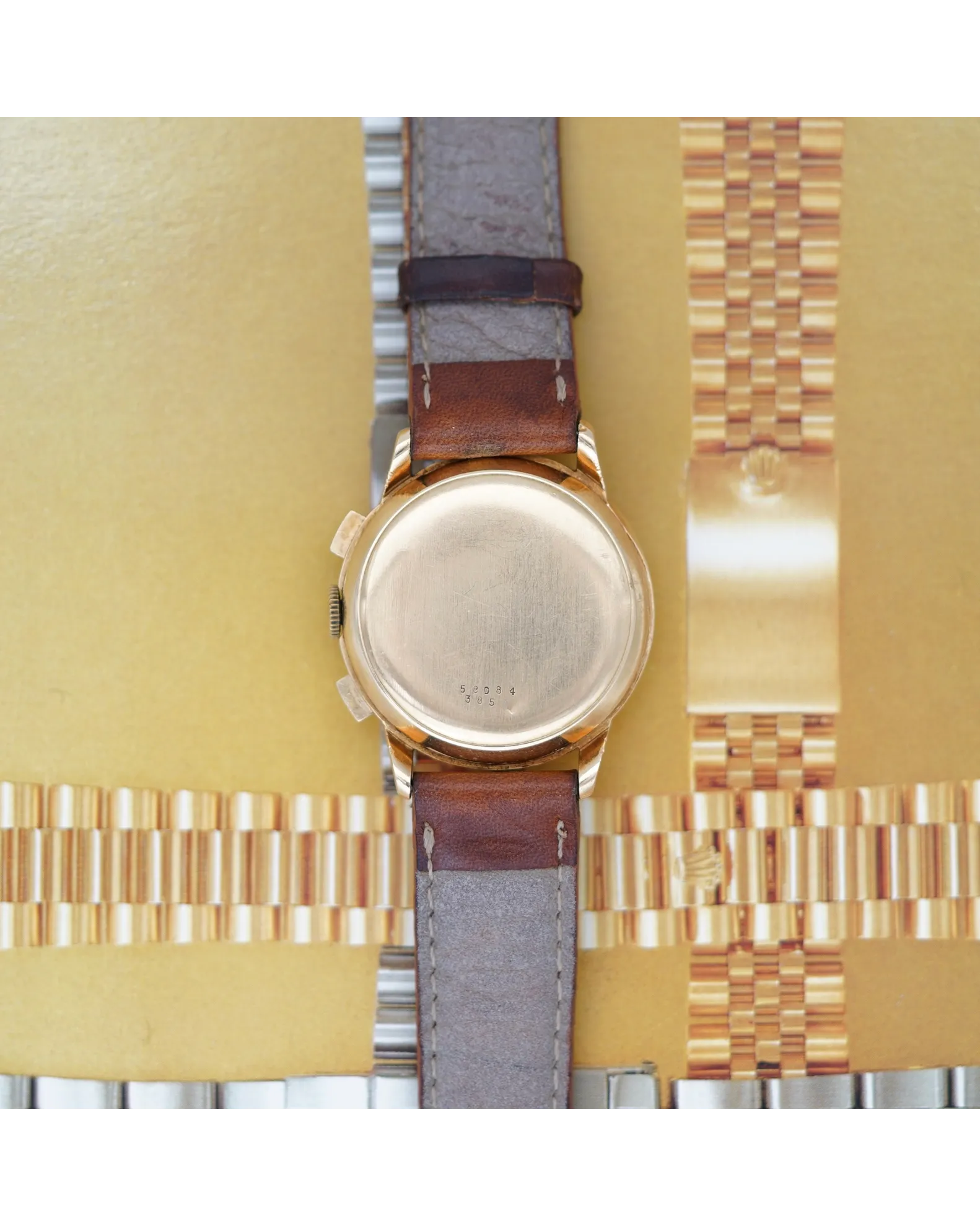 Caseback view of a vintage chronograph on brown leather strap with grey suede lining beside metal bracelets.